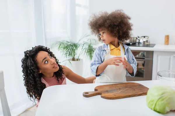 Scared african american woman looking at toddler daughter near cabbage and cutting board in kitchen — Stock Photo