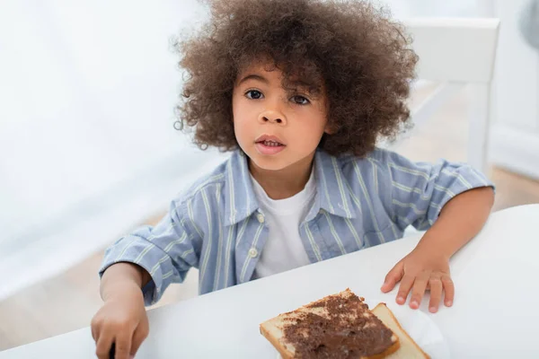 Niño afroamericano mirando la cámara cerca del pan con pasta de chocolate en la cocina - foto de stock