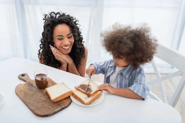 Sorrindo afro-americana mãe olhando para a criança espalhando pasta de chocolate no pão na cozinha — Fotografia de Stock