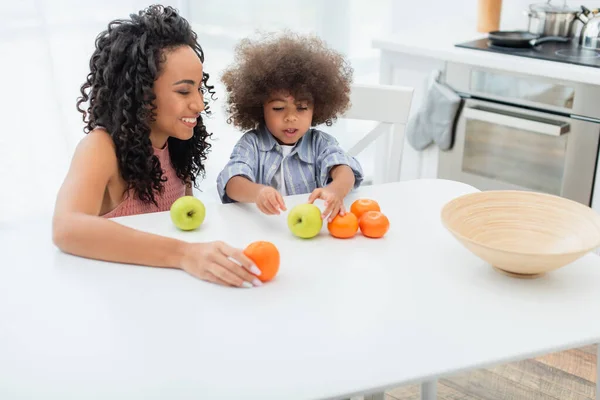 Smiling african american mom holding orange near daughter in kitchen — Stock Photo