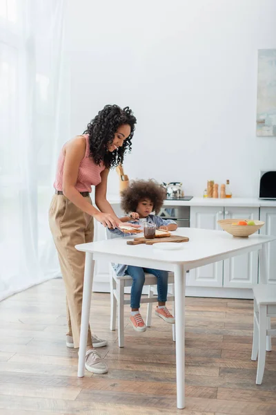 Junge Afroamerikanerin verteilt Schokoladenpaste auf Brot in der Nähe von Kindern und Früchten in der Küche — Stockfoto