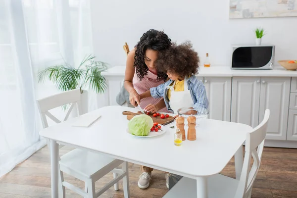 Família afro-americana cortando tomates cereja perto de óleo e repolho na cozinha — Fotografia de Stock