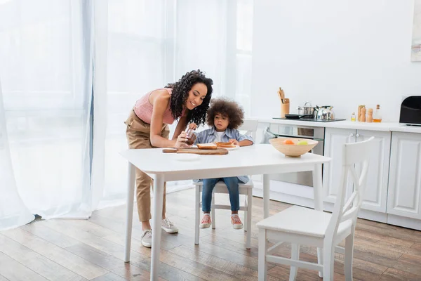 Sonriente mamá afroamericana sosteniendo pasta de chocolate cerca de su hija, pan y frutas en la cocina - foto de stock