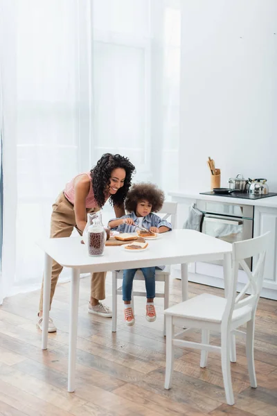 Sonriente mamá afroamericana mirando a los niños esparciendo pasta de chocolate en el pan y los cereales en casa - foto de stock