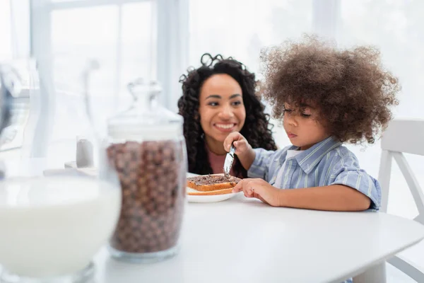 Toddler african american kid spreading chocolate paste on bread near blurred mom in kitchen — Stock Photo