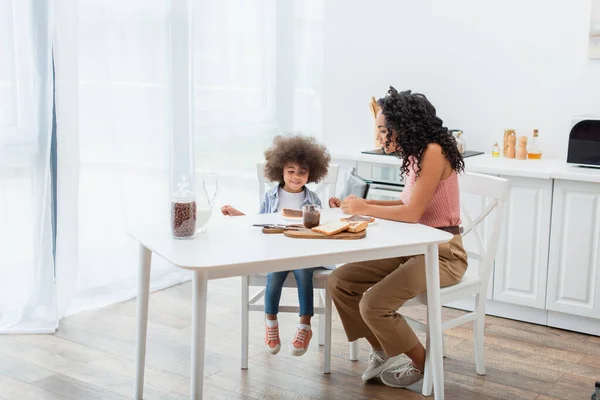 Africano americano mamá y niño sentado cerca del desayuno y la leche en la cocina - foto de stock