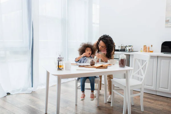 African american mom feeding daughter near breakfast and drinks in kitchen — Stock Photo