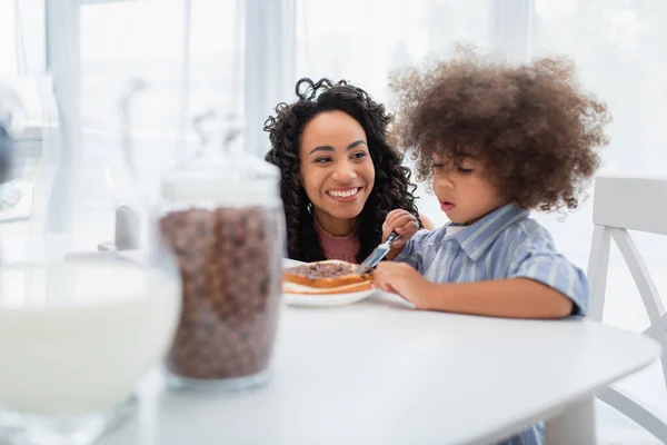 Smiling african american mom looking at blurred daughter spreading chocolate paste on bread in kitchen — Stock Photo