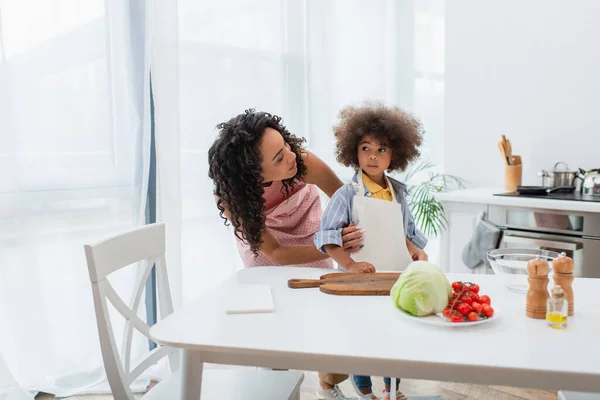 Afrikanisch-amerikanischer Elternteil umarmt Tochter in Schürze neben Gemüse in Küche — Stockfoto