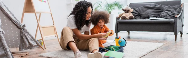 Positive afrikanisch-amerikanische Mutter hält Notizbuch in der Nähe des Rucksacks, Globus und Farbstifte zu Hause, Banner — Stockfoto