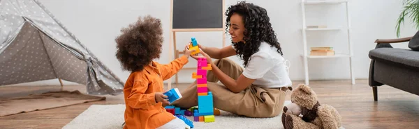 Sorrindo afro-americana mãe jogando blocos de construção coloridos perto de teepee em casa, banner — Fotografia de Stock