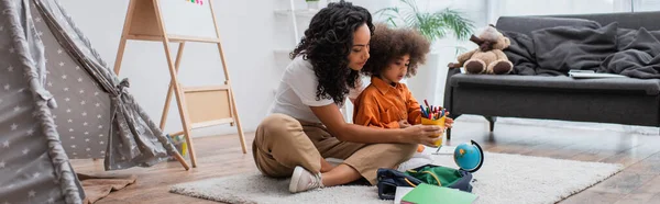 Junge Afroamerikanerin hält Buntstifte in der Nähe von Rucksack, Globus und Zelt zu Hause, Banner — Stockfoto