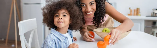 Positiva mujer afroamericana sosteniendo naranja cerca de la hija mirando a la cámara en la cocina, pancarta - foto de stock