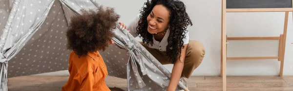 Sorrindo afro-americana mãe olhando para a filha em teepee em casa, banner — Fotografia de Stock