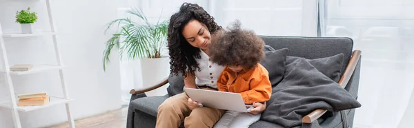 Mère afro-américaine souriante tenant un ordinateur portable près d'un enfant sur le canapé à la maison, bannière — Photo de stock