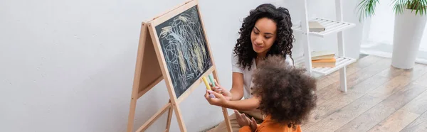 Sorridente madre afroamericana che tiene i gessetti vicino a figlia e lavagna a casa, striscione — Foto stock