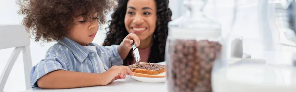 Позитивная африканская американская мама смотрит на маленькую дочь, разбрасывающую шоколадную пасту на хлеб, баннер — стоковое фото