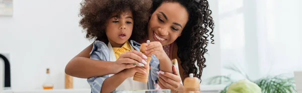 Mère afro-américaine souriante tenant moulin à poivre près de la fille dans la cuisine, bannière — Photo de stock