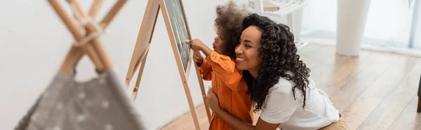 Smiling african american mom hugging daughter drawing on chalkboard near teepee at home, banner — Stock Photo