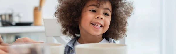 Sonriente niño afroamericano sentado cerca de cuencos y madre en la cocina, pancarta - foto de stock