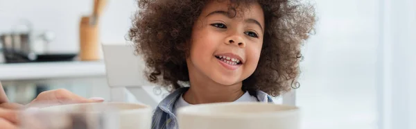 Lächelndes afrikanisch-amerikanisches Kind sitzt neben Schüsseln und Mutter in der Küche, Banner — Stockfoto