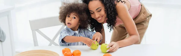 Африканская американская мама держит яблоко рядом с дочерью и апельсины на кухне, баннер — стоковое фото