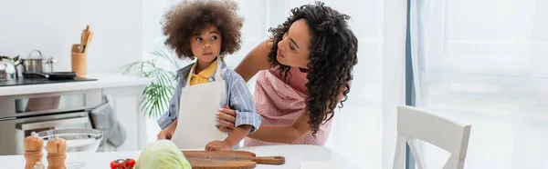 Afro-americano mãe abraçando garoto em avental perto de legumes e tigela na cozinha, banner — Fotografia de Stock