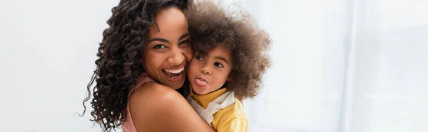 Alegre africana americana mujer abrazando hija sobresaliendo lengua en casa, pancarta - foto de stock