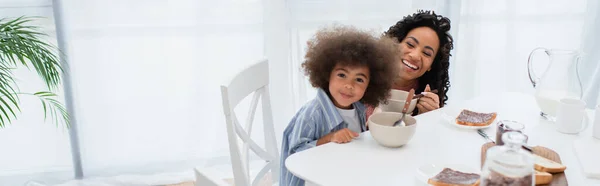 Positivo afroamericano niño mirando a la cámara cerca de mamá y sabroso desayuno en casa, pancarta - foto de stock