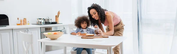 Mujer afroamericana feliz sosteniendo pasta de chocolate cerca de niño y pan en la cocina, pancarta - foto de stock
