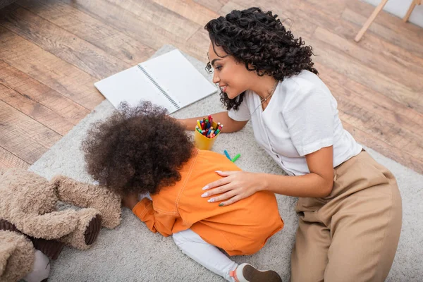 Vue grand angle de la mère afro-américaine souriante couchée près de l'enfant et des crayons de couleur à la maison — Photo de stock