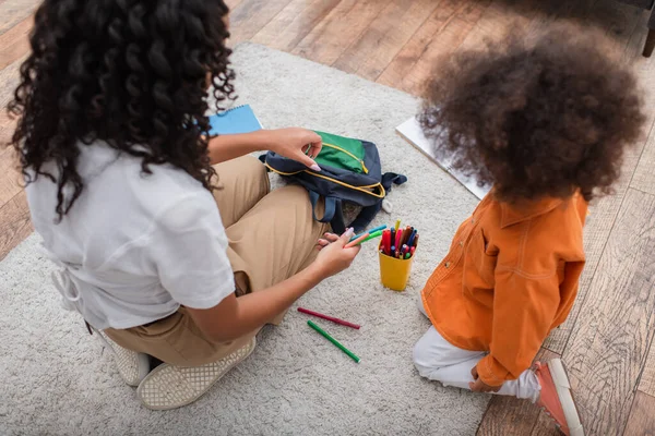 Overhead-Ansicht der afrikanisch-amerikanischen Mutter mit Buntstiften und Rucksack in der Nähe von Kind zu Hause — Stockfoto