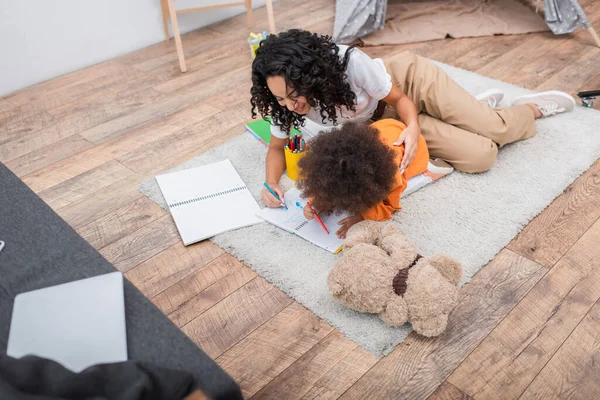 Smiling african american mom drawing on notebook near kid and teddy bear at home — Stock Photo