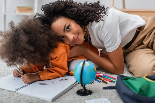 Joven afroamericana mamá mirando a la cámara cerca de dibujo infantil en el cuaderno y el globo en la alfombra - foto de stock