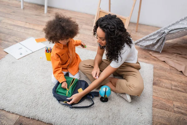 Vista de ángulo alto de madre afroamericana sosteniendo mochila cerca de niño y globo en casa - foto de stock