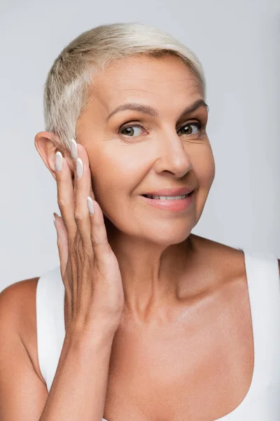 Happy senior woman smiling while applying face cream isolated on grey — Stock Photo