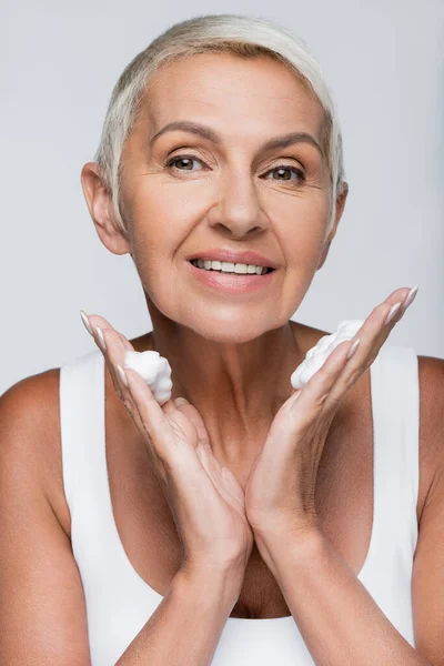 Joyeuse femme âgée avec mousse nettoyante sur les mains en regardant la caméra isolée sur gris — Photo de stock