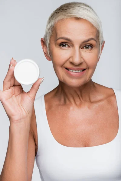 Glückliche Seniorin hält Container mit Gesichtscreme isoliert auf grau — Stockfoto