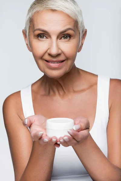 Fröhliche Seniorin hält Container mit Gesichtscreme isoliert auf grau — Stockfoto