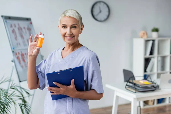 Cheerful elderly doctor holding bottle with pills and clipboard — Stock Photo