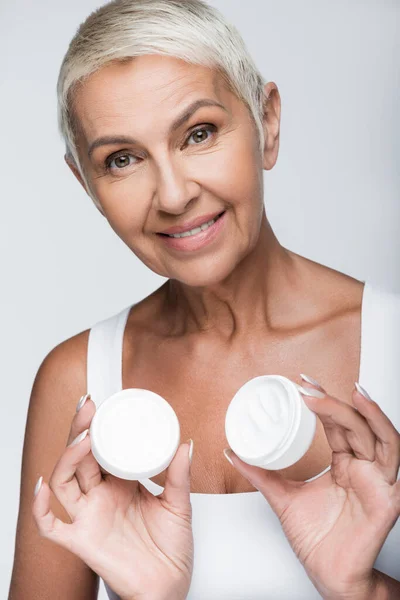 Happy senior woman holding container with cosmetic cream isolated on grey — Stock Photo