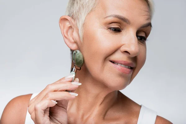 Positive senior woman massaging face with jade roller isolated on grey — Stock Photo