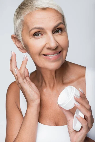 Donna anziana sorridente che tiene il contenitore con crema per il viso isolata su grigio — Foto stock