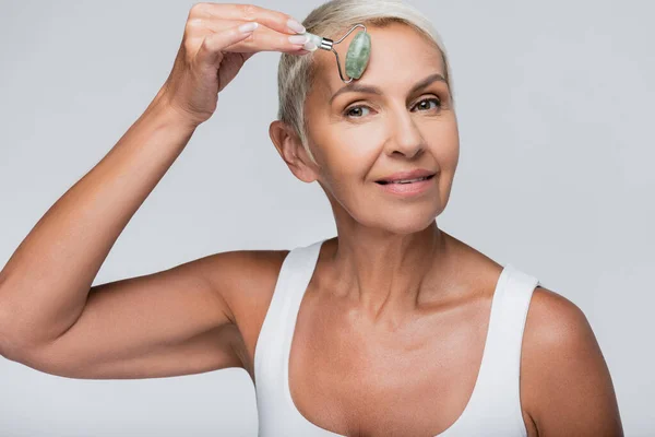 Happy senior woman massaging face with jade roller isolated on grey — Stock Photo