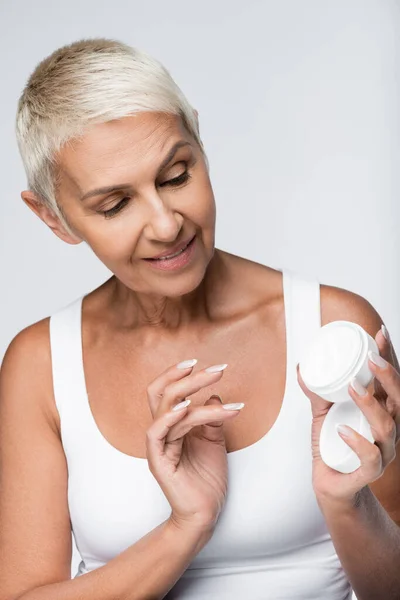 Happy senior woman looking at container with face cream isolated on grey — Stock Photo