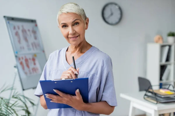 Cheerful elderly doctor holding clipboard and pen in clinic — Stock Photo