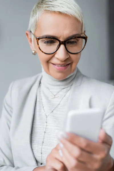 Cheerful senior businesswoman in glasses looking at smartphone — Stock Photo