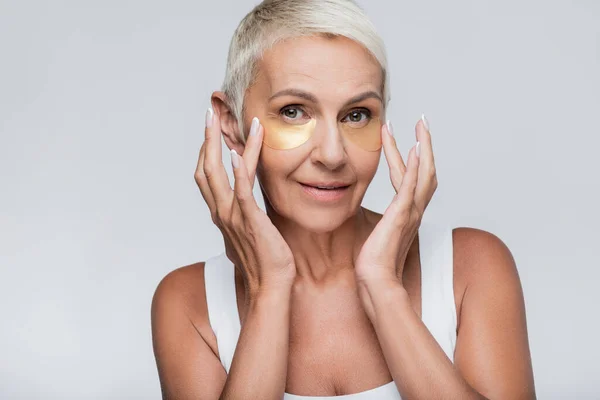 Mujer mayor satisfecha aplicando parches de colágeno aislados en gris - foto de stock