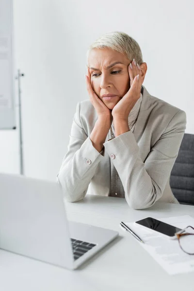 Traurige Seniorin mit Brille neben Gadgets auf dem Schreibtisch — Stockfoto