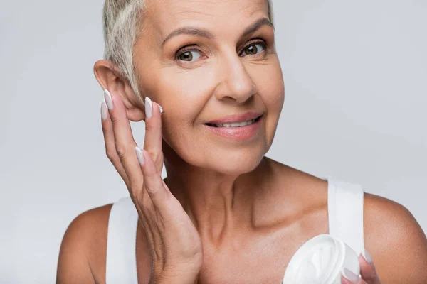 Happy senior woman holding container and applying face cream isolated on grey — Stock Photo